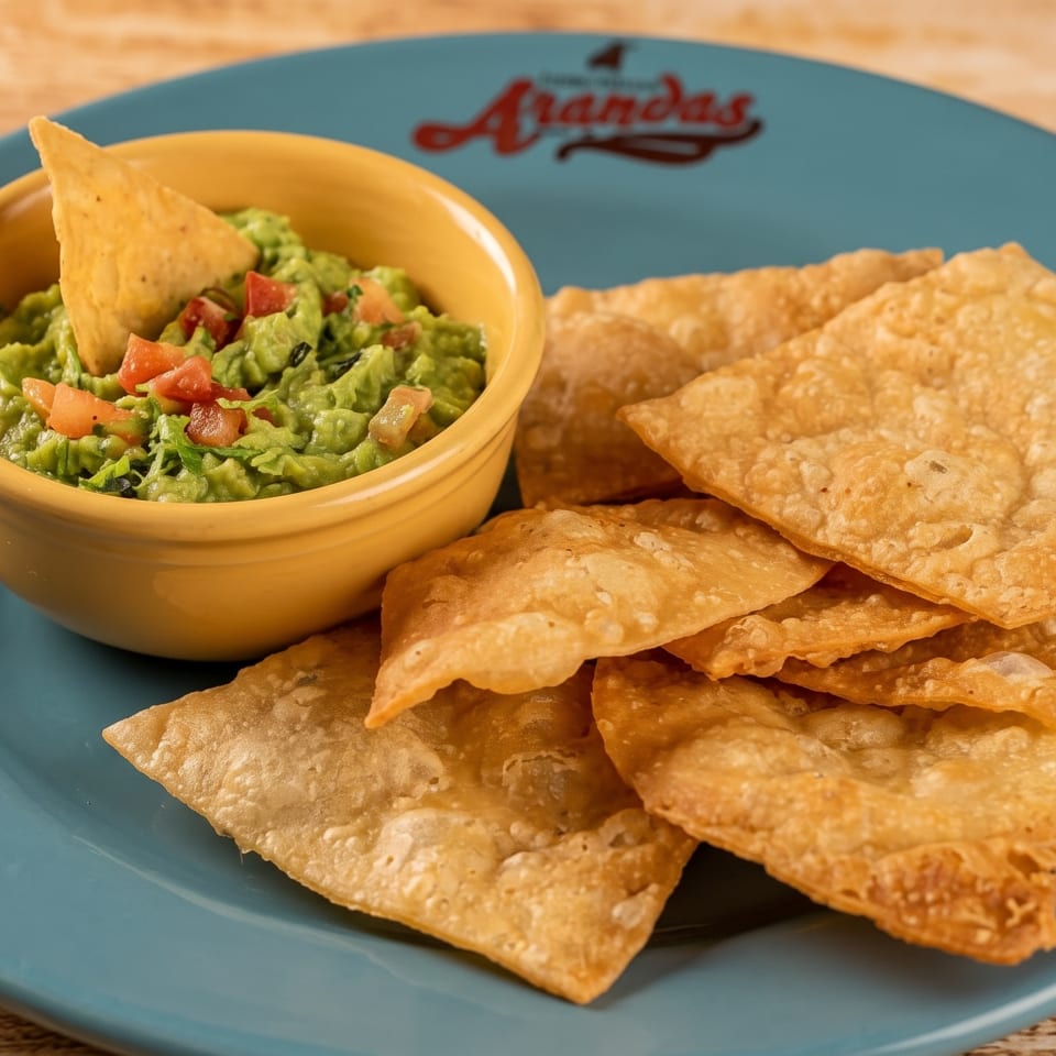 Guacamole with pico de gallo and a tortilla chip in a yellow bowl, served with fried tortilla squares on a blue plate.
