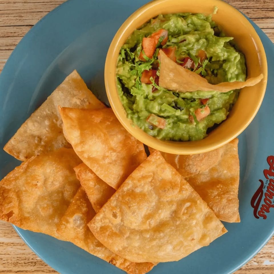 Crispy tortilla chips surround a bowl of chunky guacamole topped with diced tomatoes and cilantro on a blue plate.