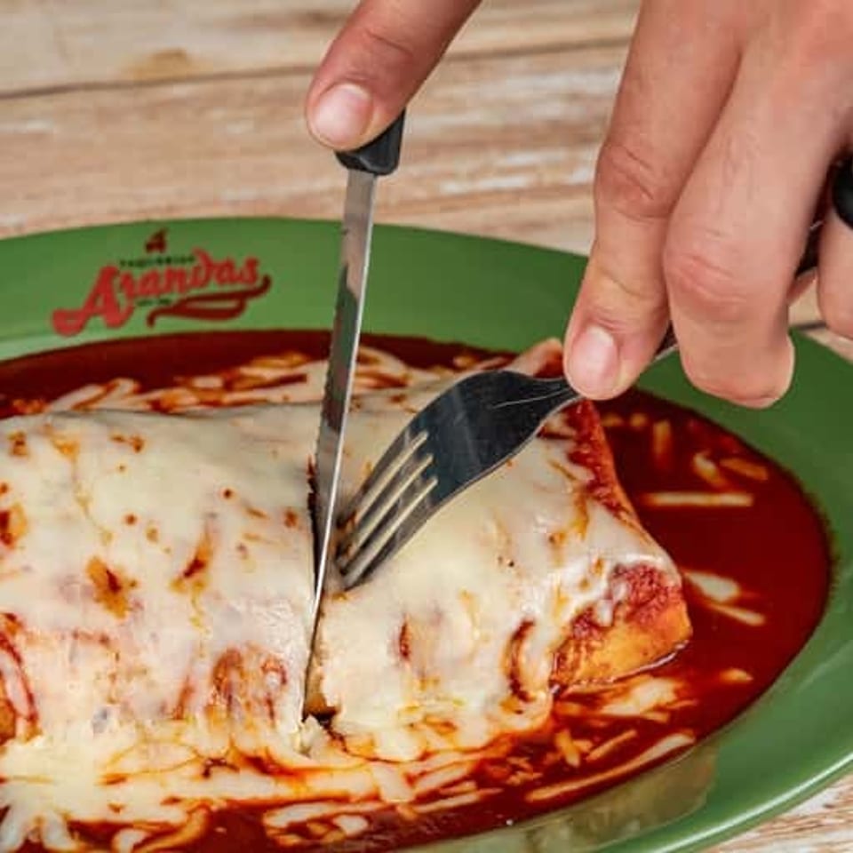 Hands cut a cheese-covered enchilada on a green plate with red sauce at a Mexican restaurant.