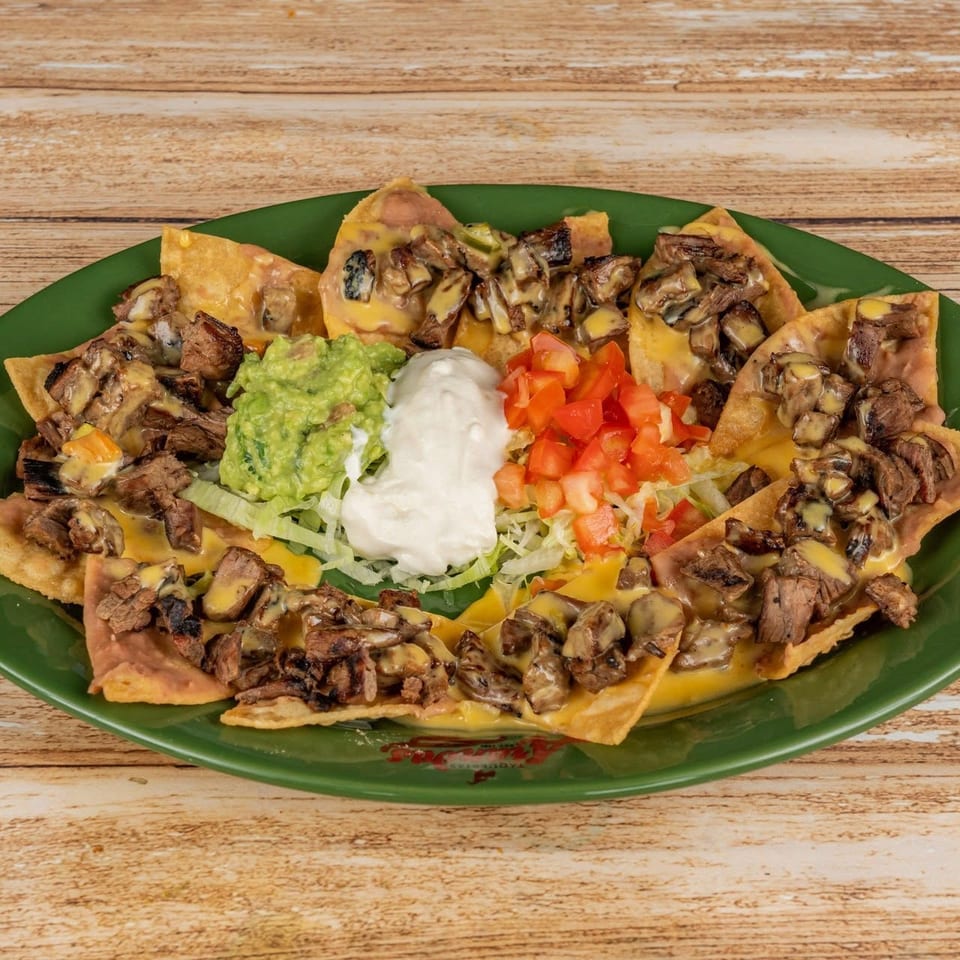 Loaded nachos topped with grilled steak, melted cheese, guacamole, sour cream, diced tomatoes, and lettuce on a green plate.