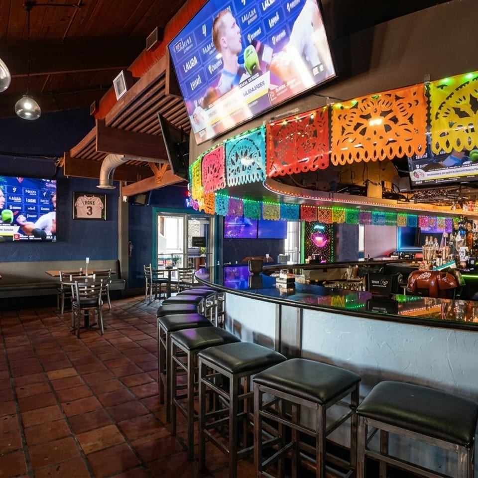 Vibrant Mexican restaurant interior with a lively bar, colorful papel picado, and multiple sports screens.