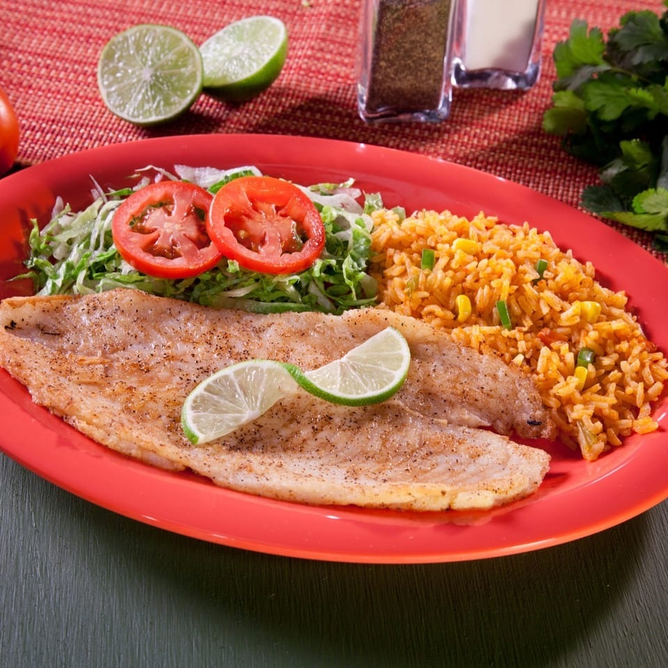 Seasoned grilled fish fillet with lime wedges, Mexican rice, and tomato-topped lettuce salad on a red plate.