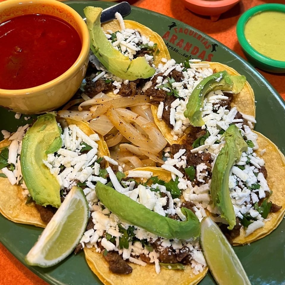Three beef tacos topped with avocado, onions, cilantro, and crumbled cheese, served with lime wedges and bowls of salsa.
