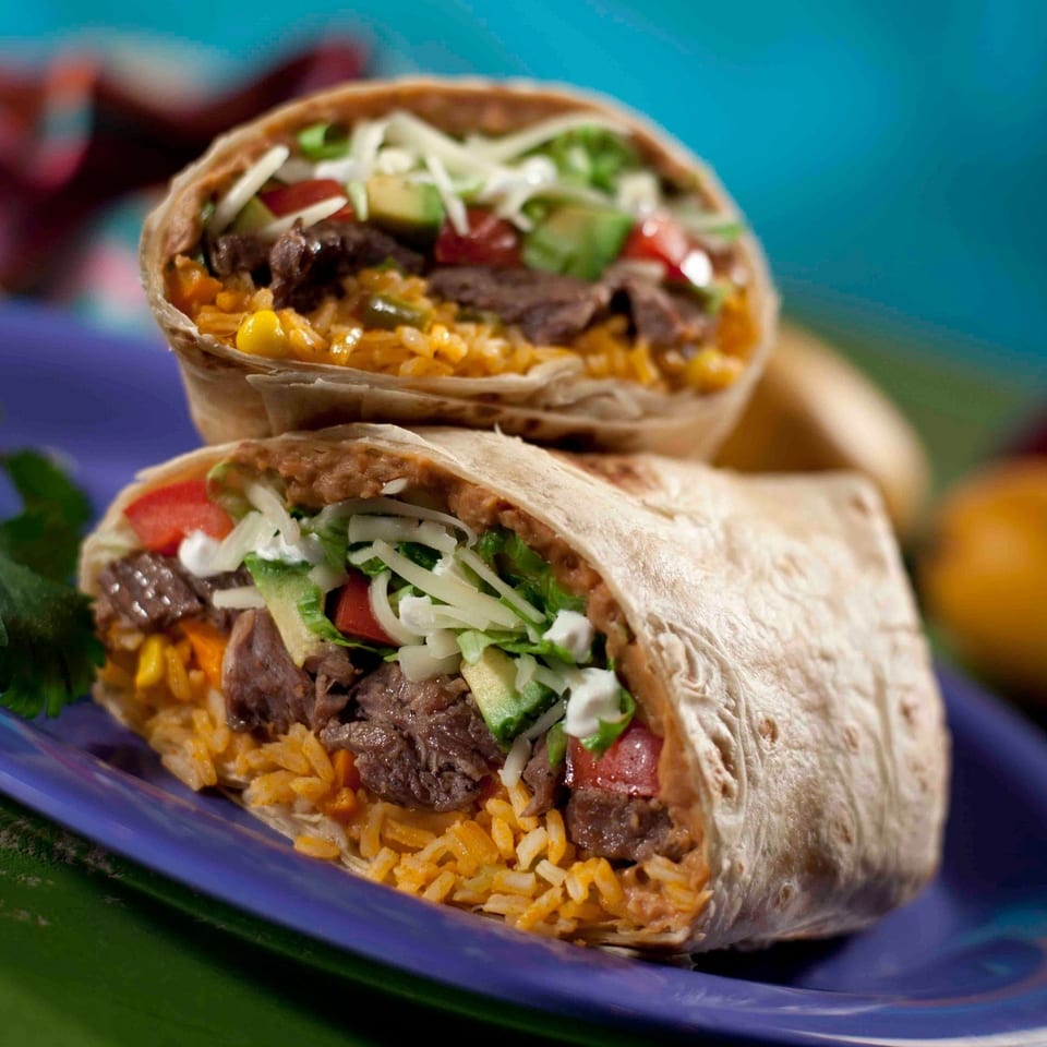 Two steak burritos filled with seasoned rice, beans, lettuce, tomatoes, avocado, and shredded cheese on a blue plate.